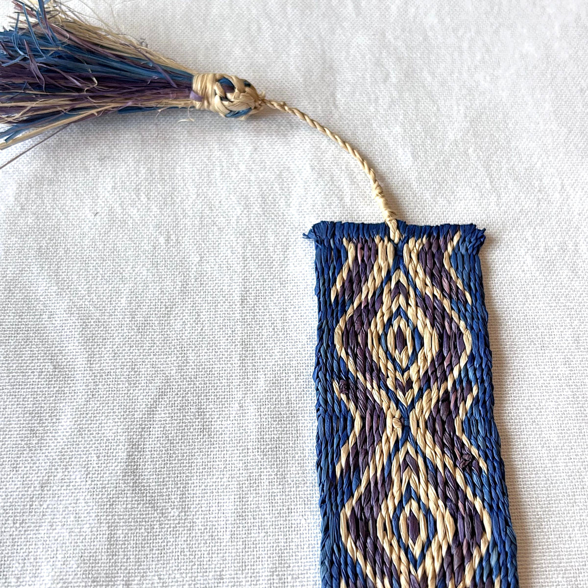 Handwoven bookmark with geometric blue, purple, and light tan pattern and tassel on a white fabric background