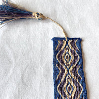 Handwoven bookmark with geometric blue, purple, and light tan pattern and tassel on a white fabric background