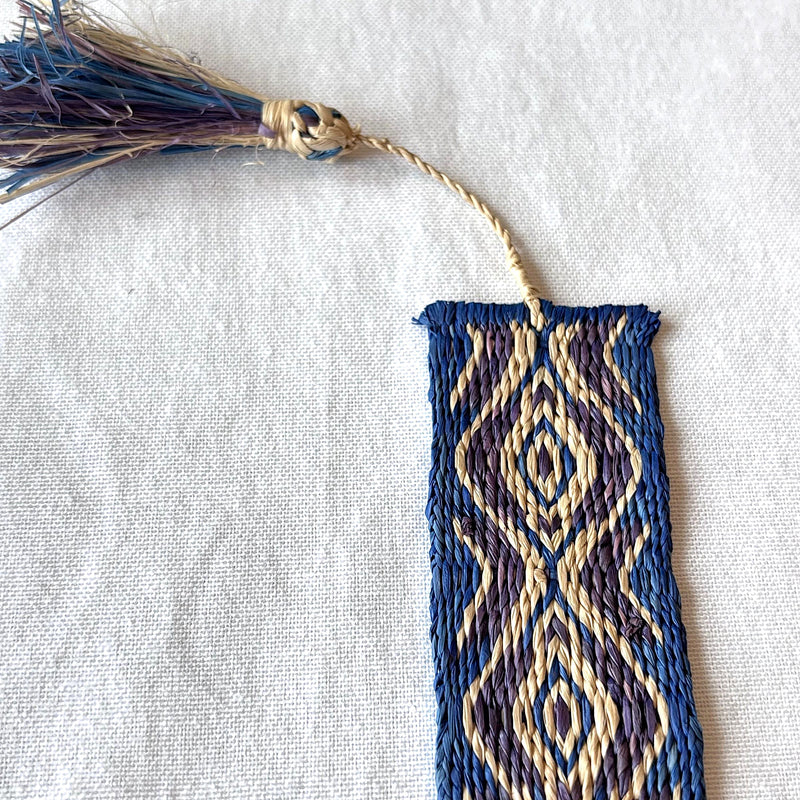 Handwoven bookmark with geometric blue, purple, and light tan pattern and tassel on a white fabric background