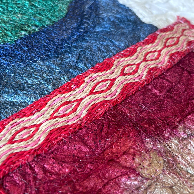 Close-up texture of red, blue, and gold material.  Colorful collaged textile artwork with nature-inspired geometric patterns in gold, white, blue, green, and red.  Hand-crafted, fair trade certified, sustainably sourced raw materials.  Made from Madagascar silk cocoons and cardwoven raffia.