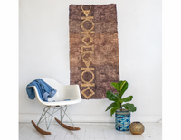A vertical brown handcrafted wild silk art piece with a gold geometric pattern hangs on a white wall in a styled room scene,.